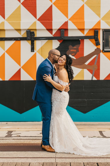 A bride and groom stand and embrace. The groom is kissing the bride on the cheek.