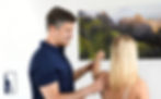 Mann in blauem Hemd prüft den Arm einer Frau in heller Umgebung. Im Hintergrund hängt ein Landschaftsbild mit grünen Bergen.