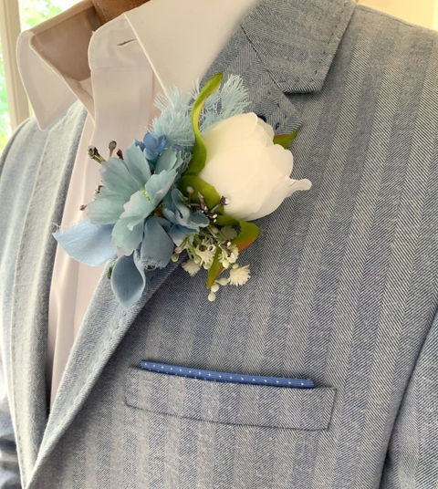 Man in Powder Blue and Pale pinstripe suit and crisp white collared shirt wearing a boutonniere featuring a pure white peony, blue organza ribbon, and blue gerbera daisy.
