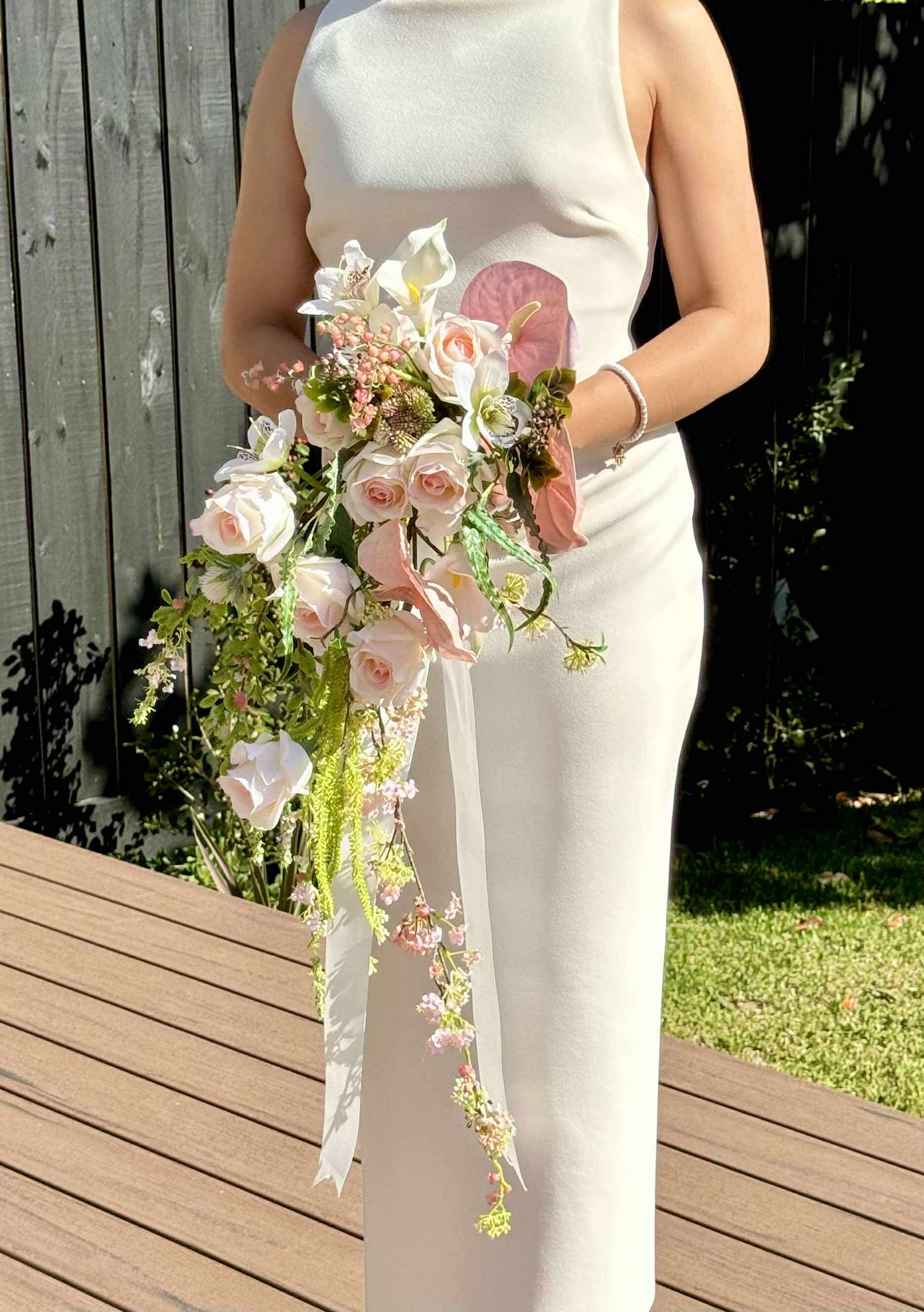 Bride in white dress holding long trailing waterfall style bouquet with soft pink roses, foliage, anthurium, white calla lily
