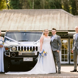 Bride, Groom, Bridesmaid and Best Man outside venue with long hummer limo. Bride holding artificial flower blue, white, yellow, bouquet.