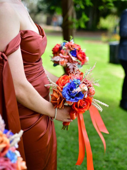 Wedding by Joy Wreaths presents this handmade rustic Australian natives bridesmaid bouquet as shown in the photographs - rust orange roses, blue lisianthus. 2 bridesmaids in a line are holding the WJW handmade bouquets, wearing copper rust off-shoulder dresses.