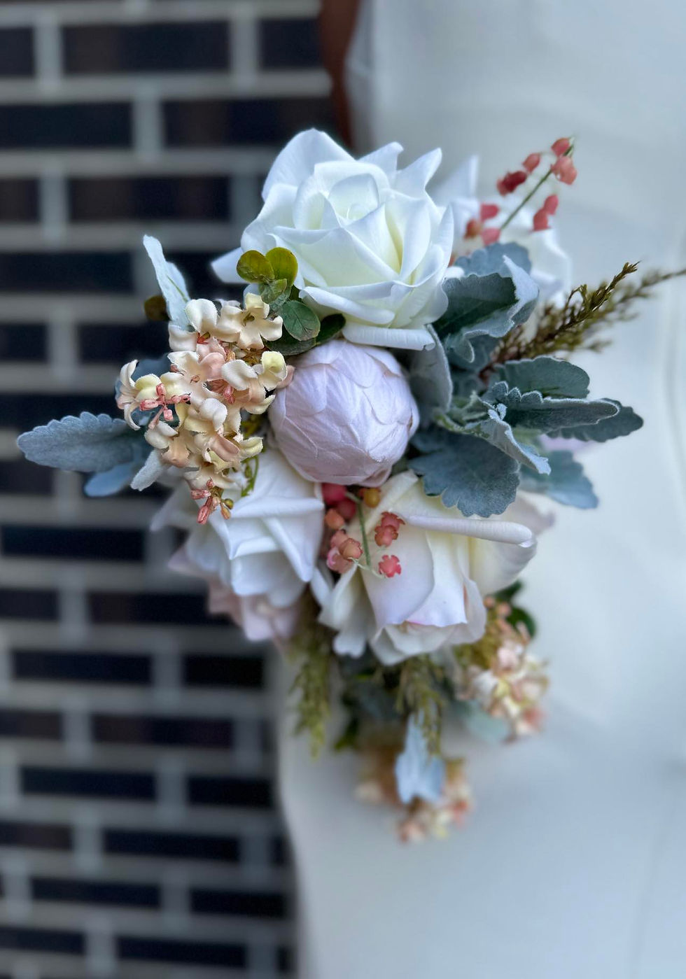 Thumbnail: Bride in white dress holding petite waterfall teardrop shape bouquet with soft pink roses, green silver suede leaves & protea