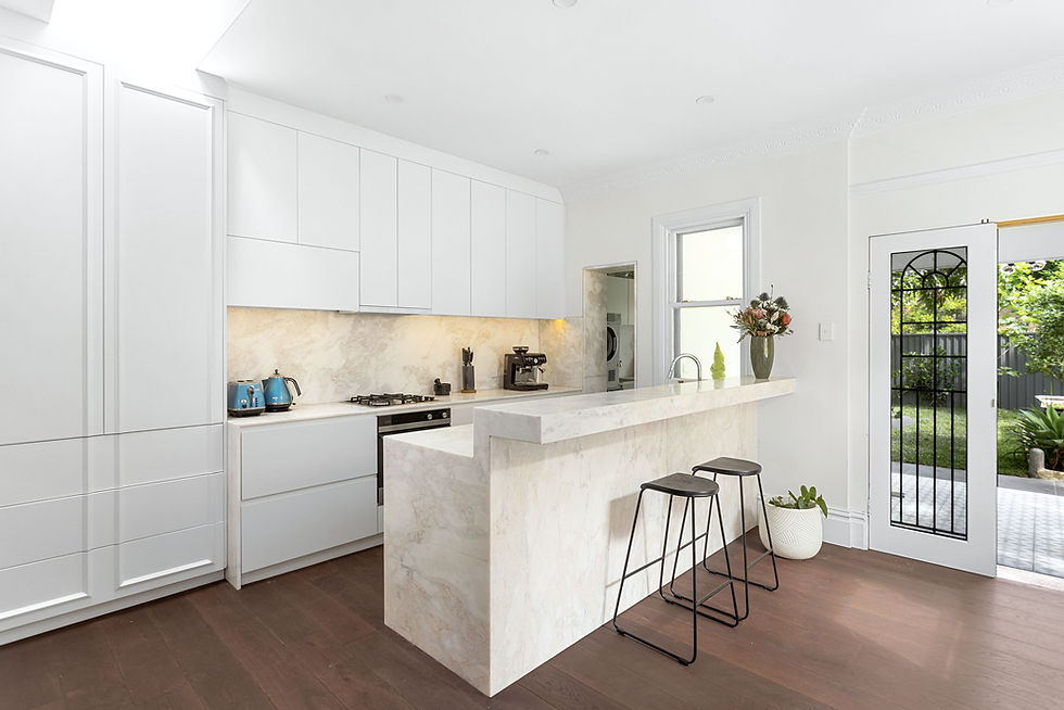 Bright kitchen with white cabinets, marble counters, blue kettle, two stools, and flowers on the bar. Door opens to a green garden.
