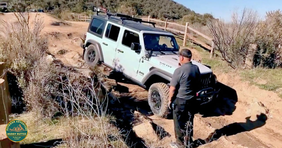 A white Jeep navigates a rocky off-road trail with a person guiding. Sunny day, grassy and hilly terrain, wooden fence in background.