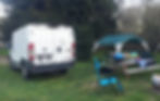 Back of van sitting on grass in campground near picnic table and sun shade.