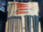 A variety of tent stakes sitting on piece of plywood. There are aluminum and steel, long stakes that look like large nails and some that look like traditional stakes.