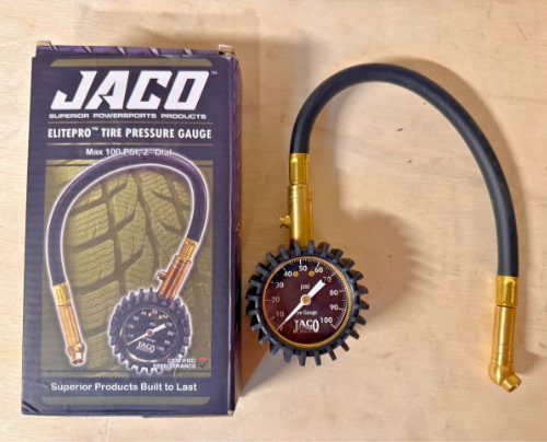 JACO tire pressure gauge next to its box on a wooden surface. Box features tire pattern and text "Superior Products Built to Last."