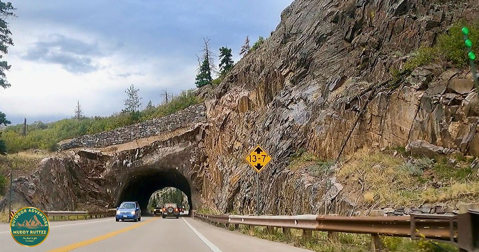 Cars drive through a rocky tunnel in a mountainous landscape. A yellow 13'-7" clearance sign is visible. Overcast sky and lush greenery.