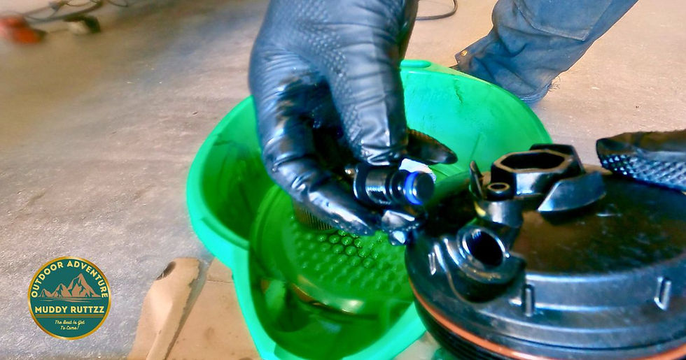 Black gloved hand is holding a Fuel Filter housing petcock is disassembled. Green drain pan on concrete floor is in background. Muddy Ruttzz Logo is visible.