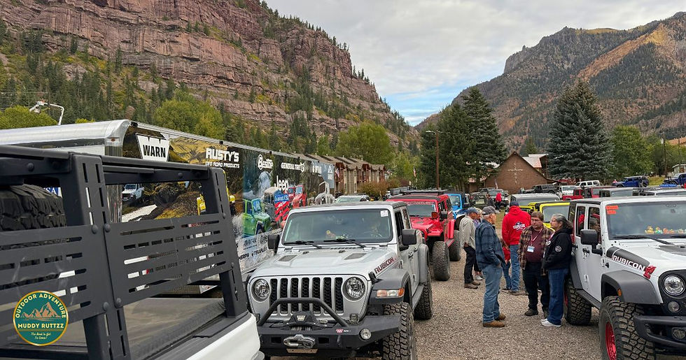 Jeeps parked with people chatting in a mountain setting. Trailers display off-road product ads. Evergreen trees and red cliffs in the background.