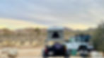 Camp spot at Joshua Tree National Park with a Exventure Off-Road Trailer. Trailer has a roof top tent in the open position and a ladder leaning on the trailer to access the tent. Jeep Wranger is sitting next to the trailer. Jeep is a Wrangler Unilimited Willys Ecodiesel. Jeep has a roof rack with traction boards mounted on top. There are Josua trees in the distance and large rocks all around.