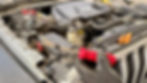 Mechanic's gloved hand holding a red tool above a Jeep engine. Nearby, orange tools rest on the engine cover. Visible "Jeep" logo.