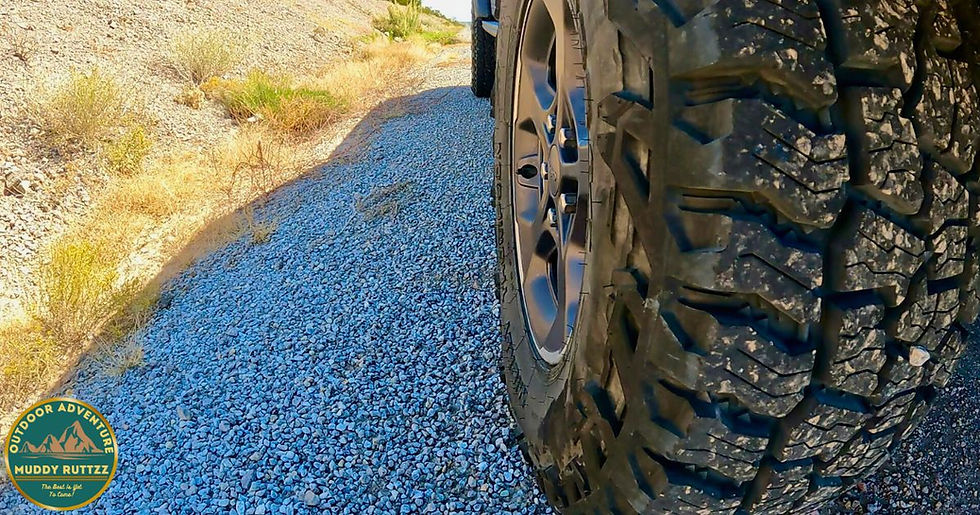 Close-up of a rugged vehicle tire on a rocky path with dry grass to the side. "Outdoor Adventure" text in corner badge. Sunny setting.