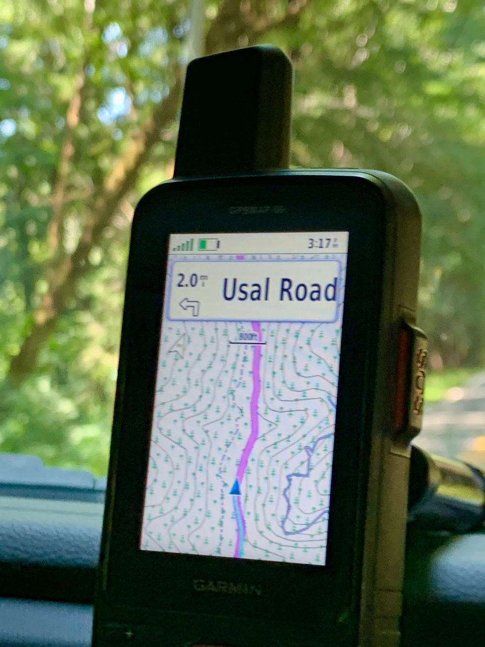GPS device displaying "Usal Road" on a map, with trees visible through a car window. The screen shows a route and distance in miles.
