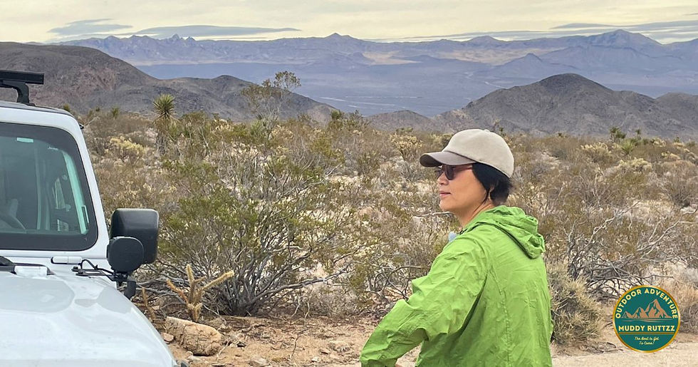 Person in a green jacket and cap stands beside a white jeep, looking at a vast desert landscape with mountains. Logo: Muddy Ruttzz Adventure.