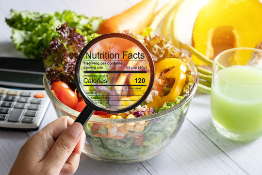 Hand holds magnifying glass over bowl of colorful salad on table, highlighting nutrition facts. Calculator and green smoothie nearby. Bright setting.