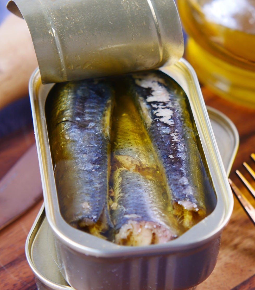 Open tin of sardines in oil on a wooden table. The fish are silvery and submerged partially. Golden fork and glass in the background.
