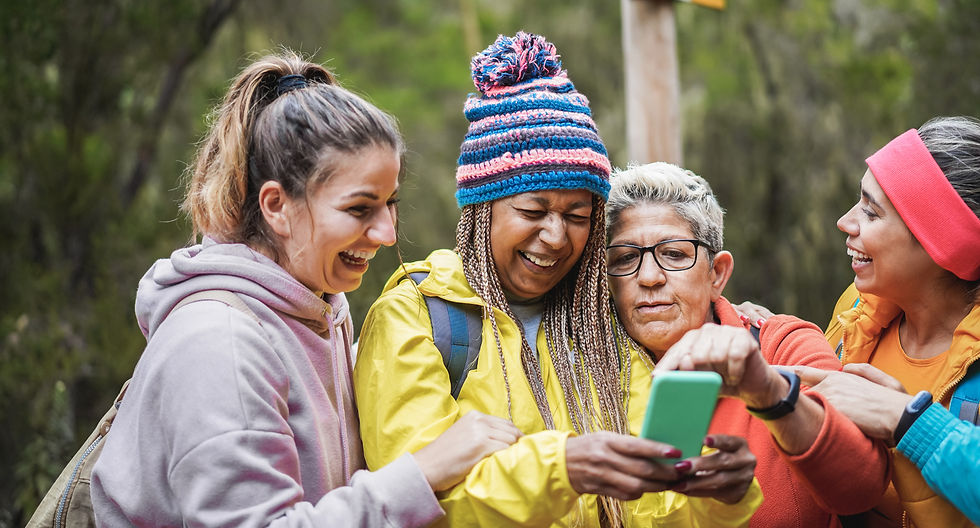 Four women in colorful outdoor gear laugh while looking at a phone. They're standing in a lush, green forest, exuding joy and camaraderie.