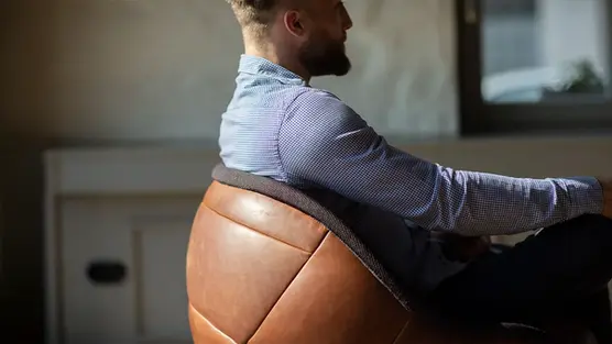 Eine Person sitzt seitlich in einem Loungesessel wembley im Retro-Fußball-Design aus braunem Vintage-Echtleder in einer Wohnatmosphäre, Hersteller lillus