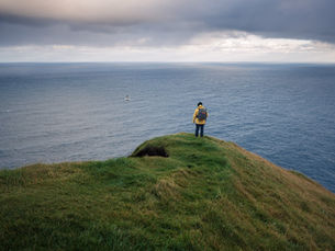 Figura con giacca gialla e zaino, ferma sulla cresta d'erba a picco sull'Atlantico. È il tema del viaggio in un fotogramma: verde e oceano, niente altro. Verticale.