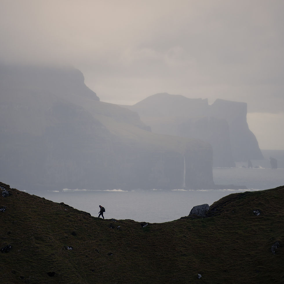Figure minuscole su collinetta verde con scogliere nella nebbia: la scala umana nel paesaggio faroese, il peso della nebbia, la solitudine fertile di chi aspetta. Rafforza il tema della resa e dell'attesa.