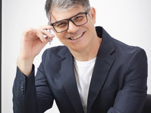 Foto do cabeleireiro Kiko Ricco, homem branco usando óculos de grau retangulares e blazer. Ele tem cabelos grisalhos estilizados. Está encostado em uma poltrona, sorrindo enquanto posa para a foto com uma tesoura para cabelos.