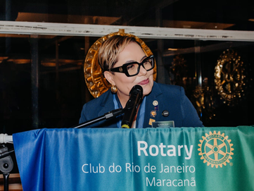 Foto da Presidente 2025-2026 Claudia Reis, muçher branca de cabelos aloirados escuros curtos, de óculos de armação grossa preta, trajando terninho azl marinho com diversos distintivos na lapela, falando a partir de tribuna de seu Clube onde se lê o nome do Clube