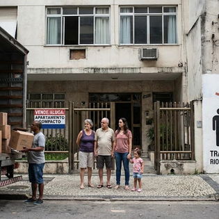 rua urbana no Rio de Janeiro. No centro, uma família composta por um casal de idosos, uma mulher jovem e uma criança pequena observa um caminhão de mudança sendo carregado. À direita, um muro branco exibe um infográfico com uma seta vermelha apontando para baixo sobre silhuetas de uma família que diminui de tamanho, acompanhado do texto: 'TRANSIÇÃO DEMOGRÁFICA RIO: MENOS PESSOAS POR LAR'. Ao fundo, um prédio residencial tem uma placa de 'Vende-se/Aluga-se: Apartamento Compacto'. O cenário inclui a clássica calçada de pedras portuguesas.