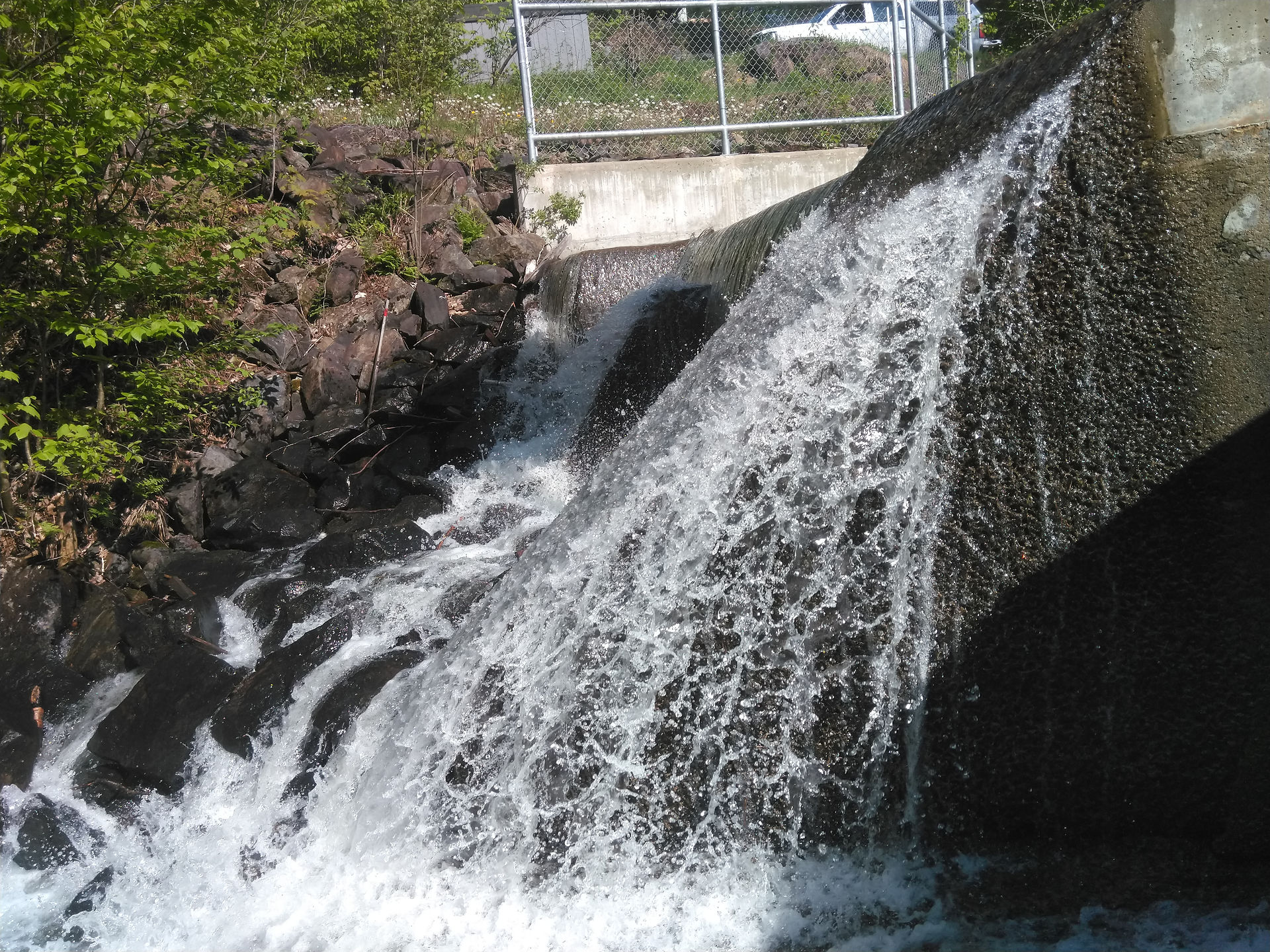 Un petit barrage en béton contrôlant le flux d’un cours d’eau à travers un dénivelé rocheux. L’eau s’écoule vigoureusement sur les rochers, créant une chute d’eau dynamique. Une clôture est visible en arrière-plan, indiquant un accès réglementé au site.