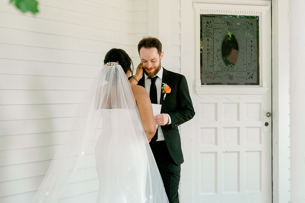 Bride in white dress touches groom's cheek as he reads a note. White wall and ornate door in background. Warm, emotional scene.
