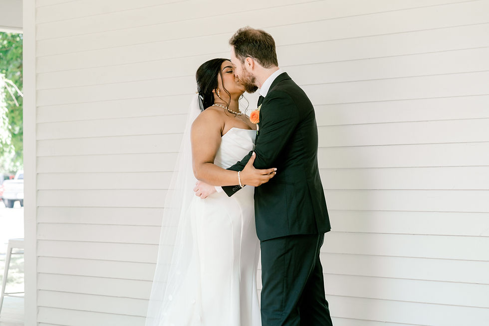 Bride in white gown and veil kisses groom in black suit with orange boutonniere, against white wooden wall background. Romantic setting.