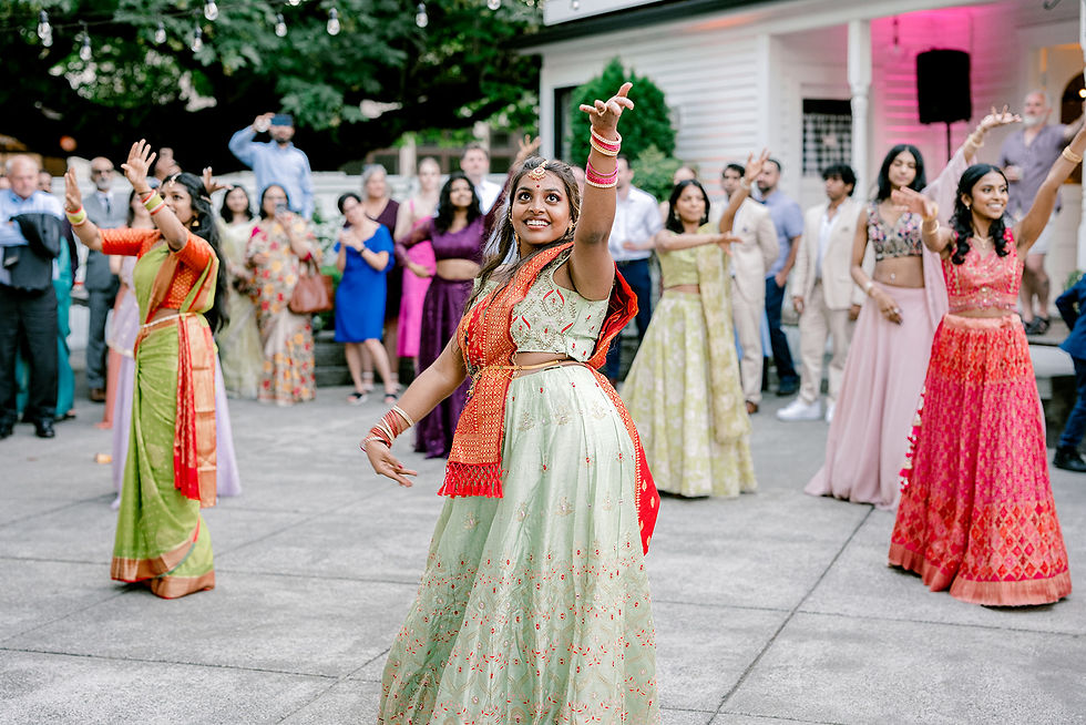 Women in colorful traditional attire dance joyfully at an outdoor event, with a crowd in the background under string lights.