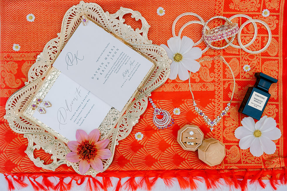 Ornate wedding invitation on orange patterned fabric with white flowers, jewelry, a perfume bottle, and rings. Bright and festive.