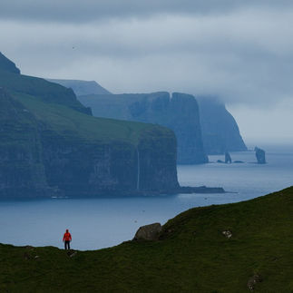 Un socio di Blu Puffin in giacca rossa ammira il paesaggio drammatico delle Isole Faroe: scogliere verdi, mare calmo e isole rocciose sotto un cielo nuvoloso. #ViaggiFotografici #Faroe #FotografiaCreativa