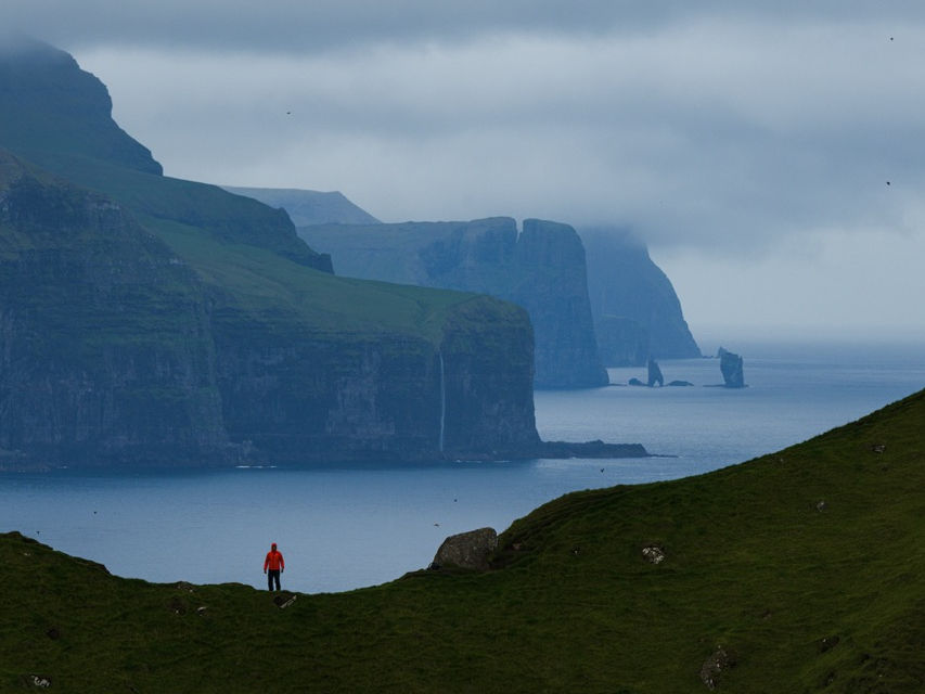 Un socio di Blu Puffin in giacca rossa ammira il paesaggio drammatico delle Isole Faroe: scogliere verdi, mare calmo e isole rocciose sotto un cielo nuvoloso. #ViaggiFotografici #Faroe #FotografiaCreativa