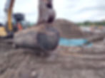 An excavator using its bucket to lift a cylindrical fuel tank out of an excavation area and over to a blue membrane to prevent cross-contamination