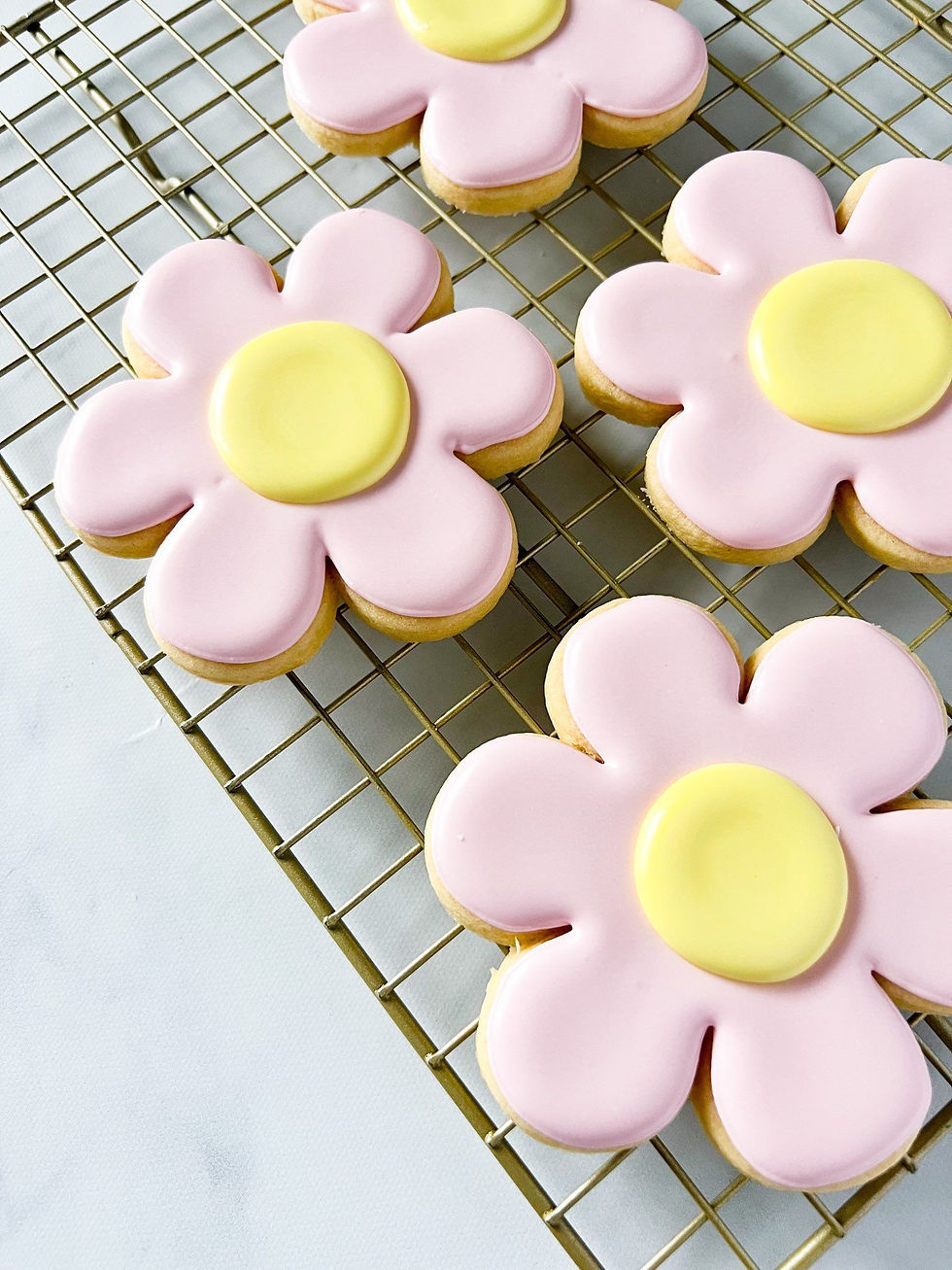 Flower-shaped sugar cookies decorated with pastel pink petals and a yellow center, arranged on a gold cooling rack. The cookies have smooth, glossy icing and a simple, cheerful design, resembling cartoon-style flowers. The background is a light, clean surface, creating a bright and inviting look.