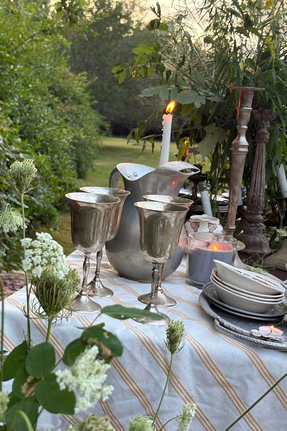 Garden table at sunset with flowers, stacked teacups, apples, candles, and bowls in a lush green outdoor setting.