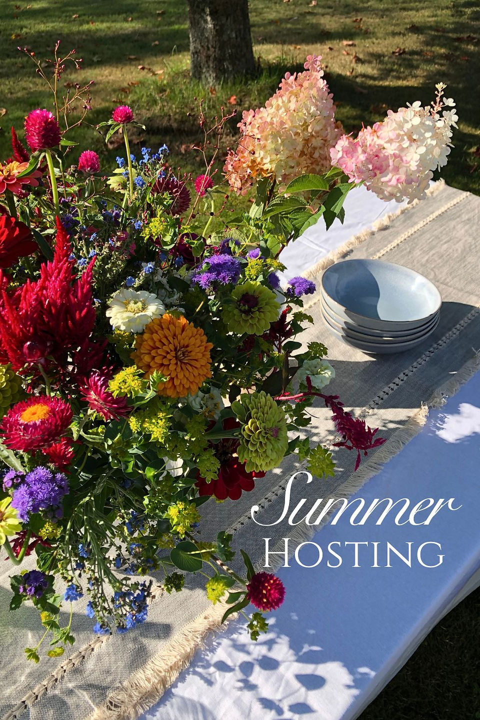 Outdoor table with colorful flower arrangements, stacked plates, bowls, and wine bottle on white cloth in a sunny picnic table. Summer Hosting text.