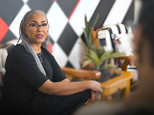 Woman with glasses and braided hair, seated in a colorful room with a geometric wall pattern, talking to an unseen person, looking composed.