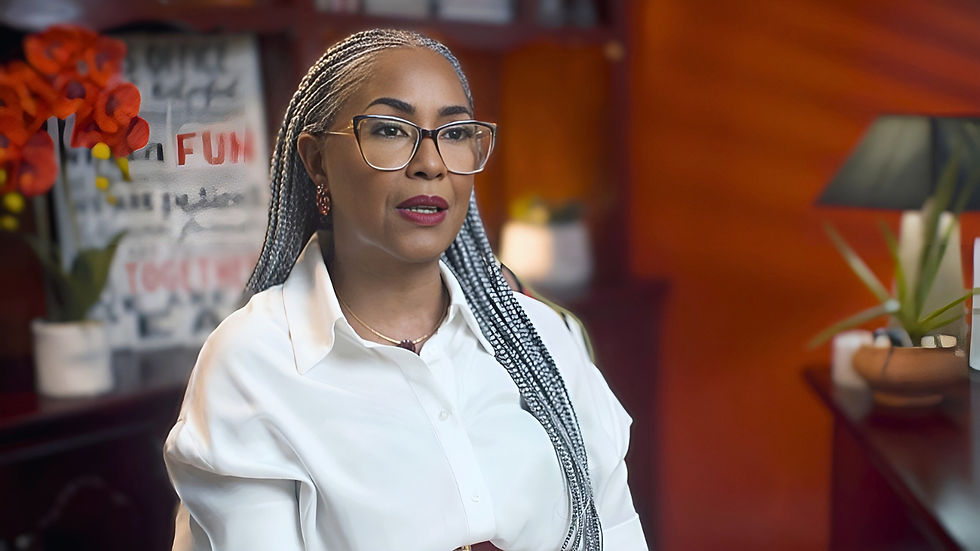 Woman with braided hair and glasses speaks in a room. White shirt contrasts orange wall. Red flowers and text art in the background.