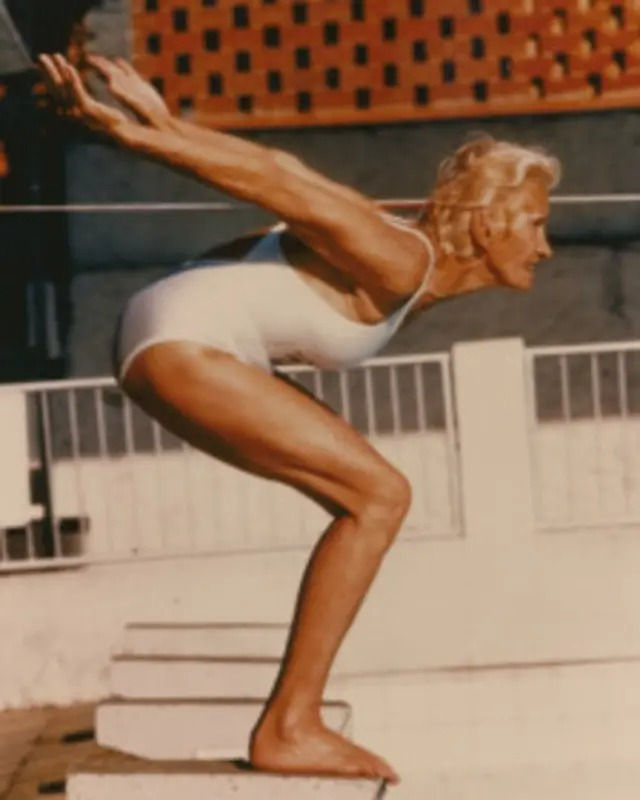 Fotografia antiga colorizada de Maria Lenk de maiô branco, em posição de mergulho sobre o bloco de largada.