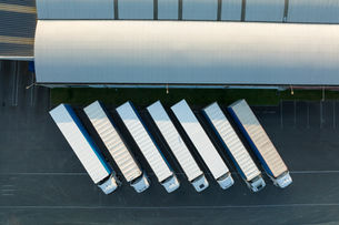 Aerial view of seven white semi-trucks lined up beside a large warehouse loading dock on dark pavement.