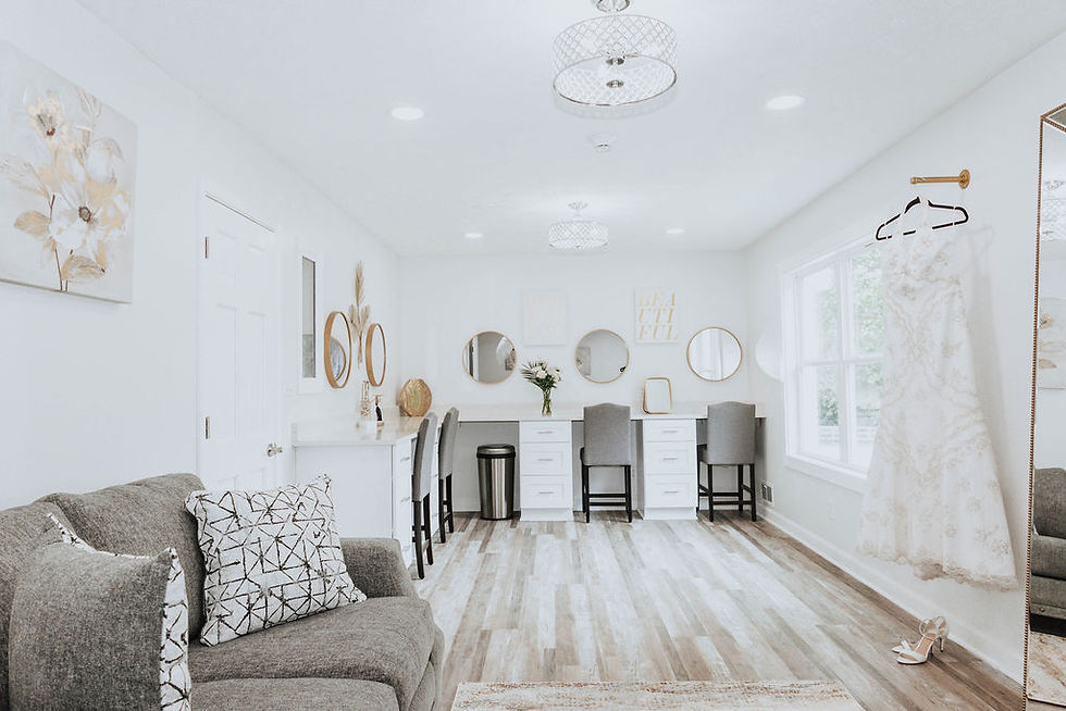Bright bridal dressing room with white walls, vanity, and gray chairs. A lace wedding gown hangs near a window. Cozy couch with patterned pillows.