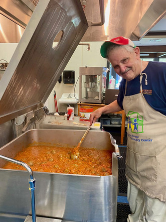 Smiling man stirring large stew pot, Food Bank of Monmouth County apron.