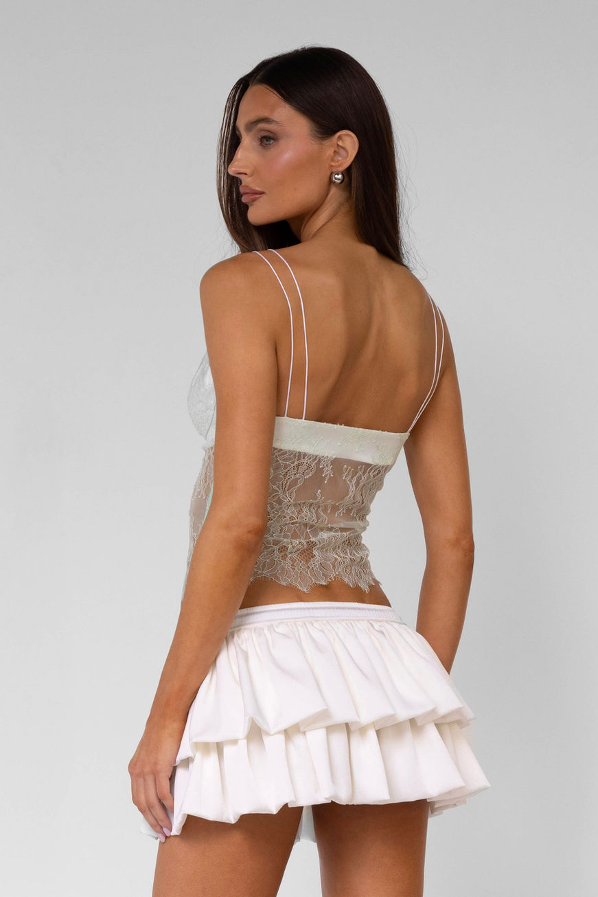 Full length shot of a model facing away from the camera and her head turned to her left, wearing a white ruffled mini skirt and a mint green lace top