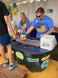 Man in "RELEASE THE DUCK!" shirt helps child at duck race game.