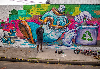 Painel do artista Pakato: seu parsonagem que é uma lata de spray cologando uma garrafa na lixeira para reciclagem.