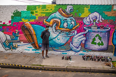 Painel do artista Pakato: seu parsonagem que é uma lata de spray cologando uma garrafa na lixeira para reciclagem.
